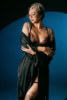A stylish woman poses in a black robe and lingerie, adorned with tattoos.