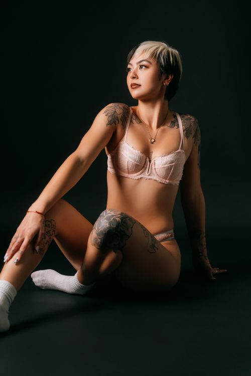 Free Tattooed woman posing in lingerie against a dark studio backdrop, expressing confidence. Stock Photo
