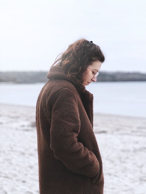 Free Brunette woman in cozy brown coat standing on beach during winter in La Forêt-Fouesnant, France. Stock Photo