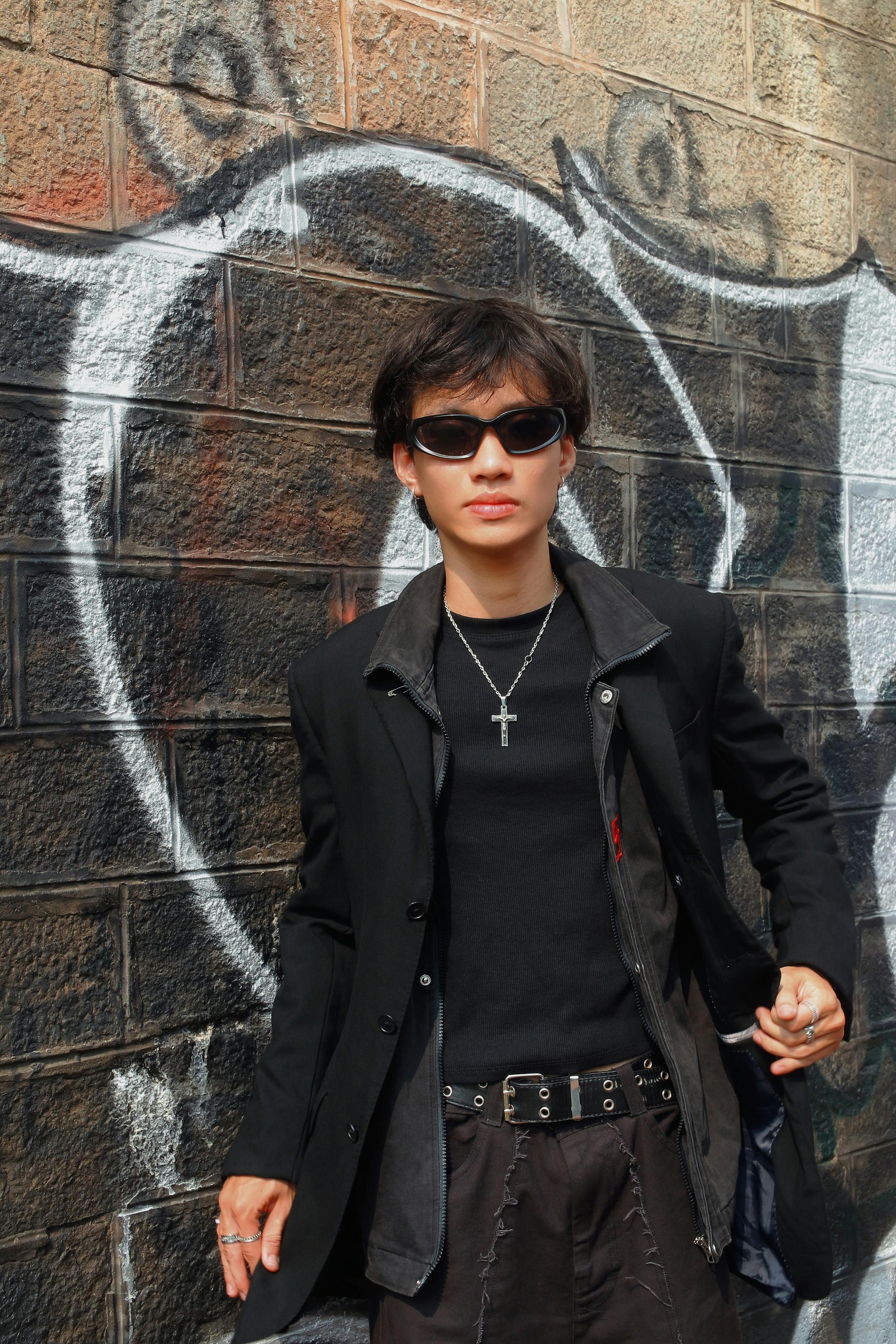 Confident young man wearing sunglasses and black outfit poses by urban graffiti wall.
