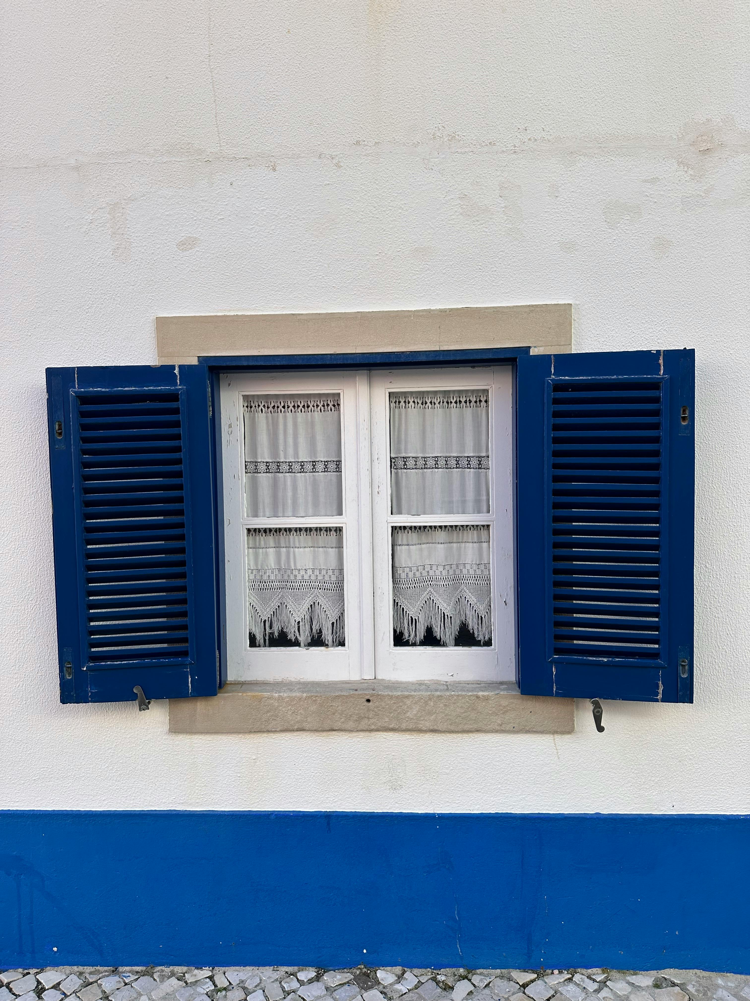 Elegant blue shuttered window on white wall in Ericeira, Portugal.