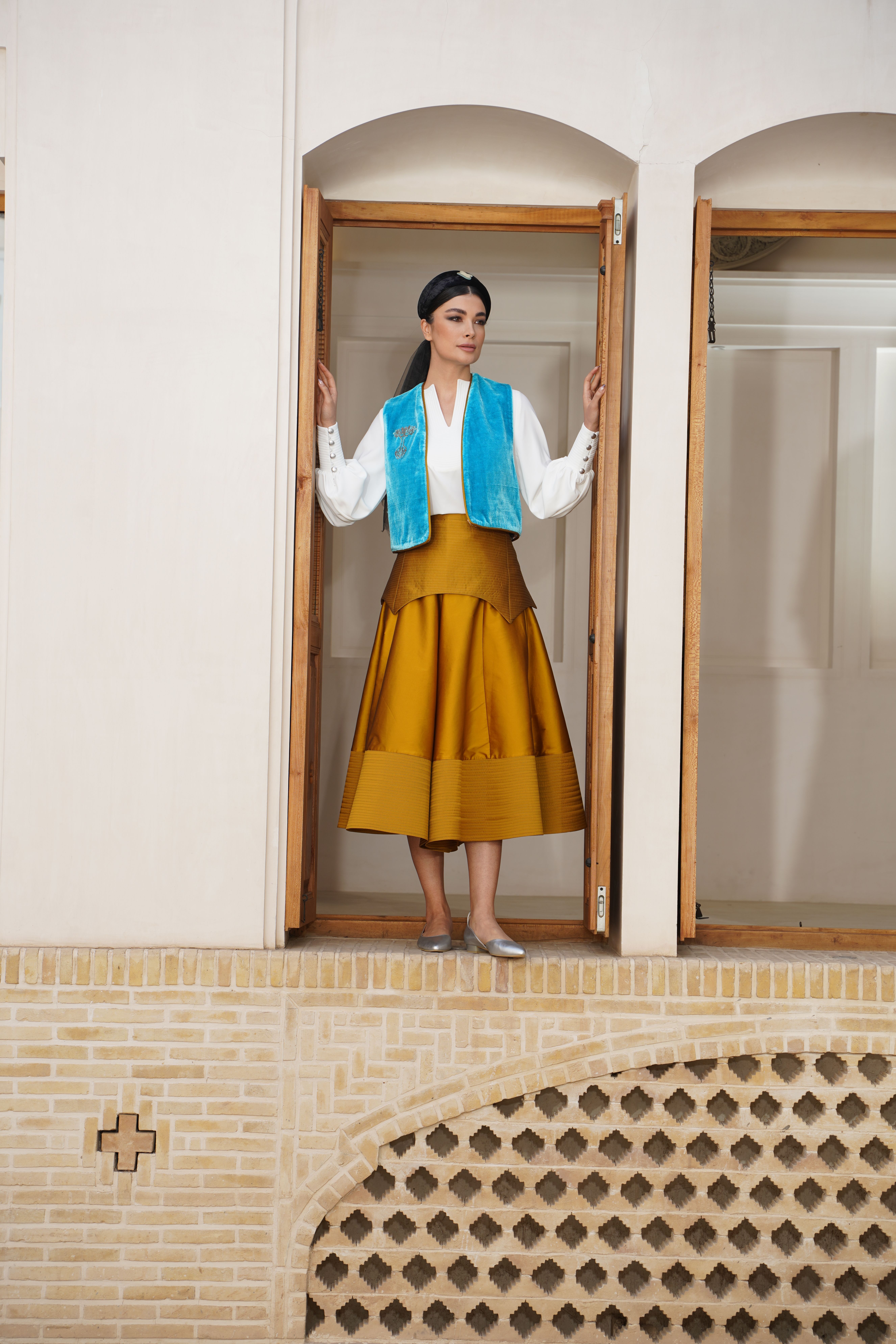 Model stands in doorway, showcasing elegance in traditional attire with blue vest and mustard skirt.