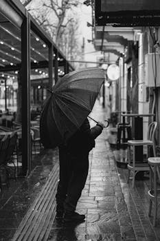 Black-and-white image of a person with umbrella on a rainy urban sidewalk.