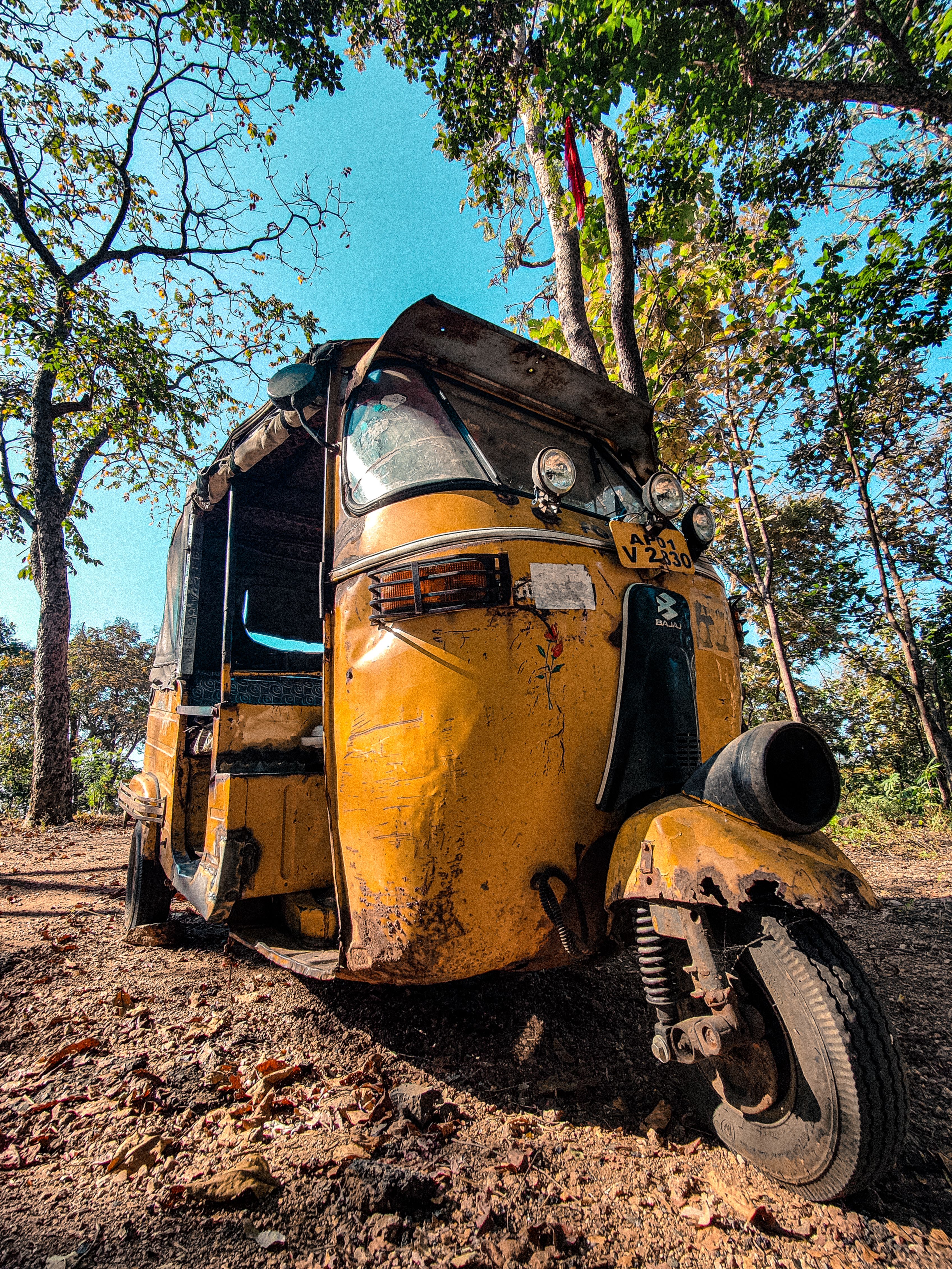 A vintage yellow auto rickshaw standing unused in a rural forest setting.