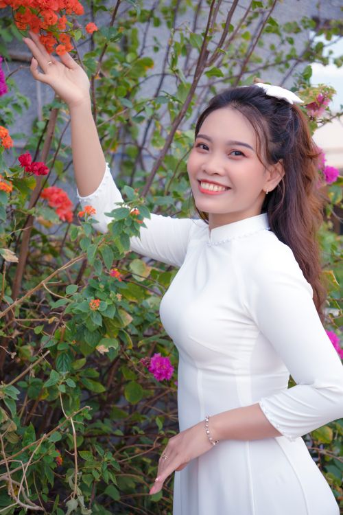 Free A young Vietnamese woman in a white ao dai smiles while standing in a vibrant flower garden. Stock Photo