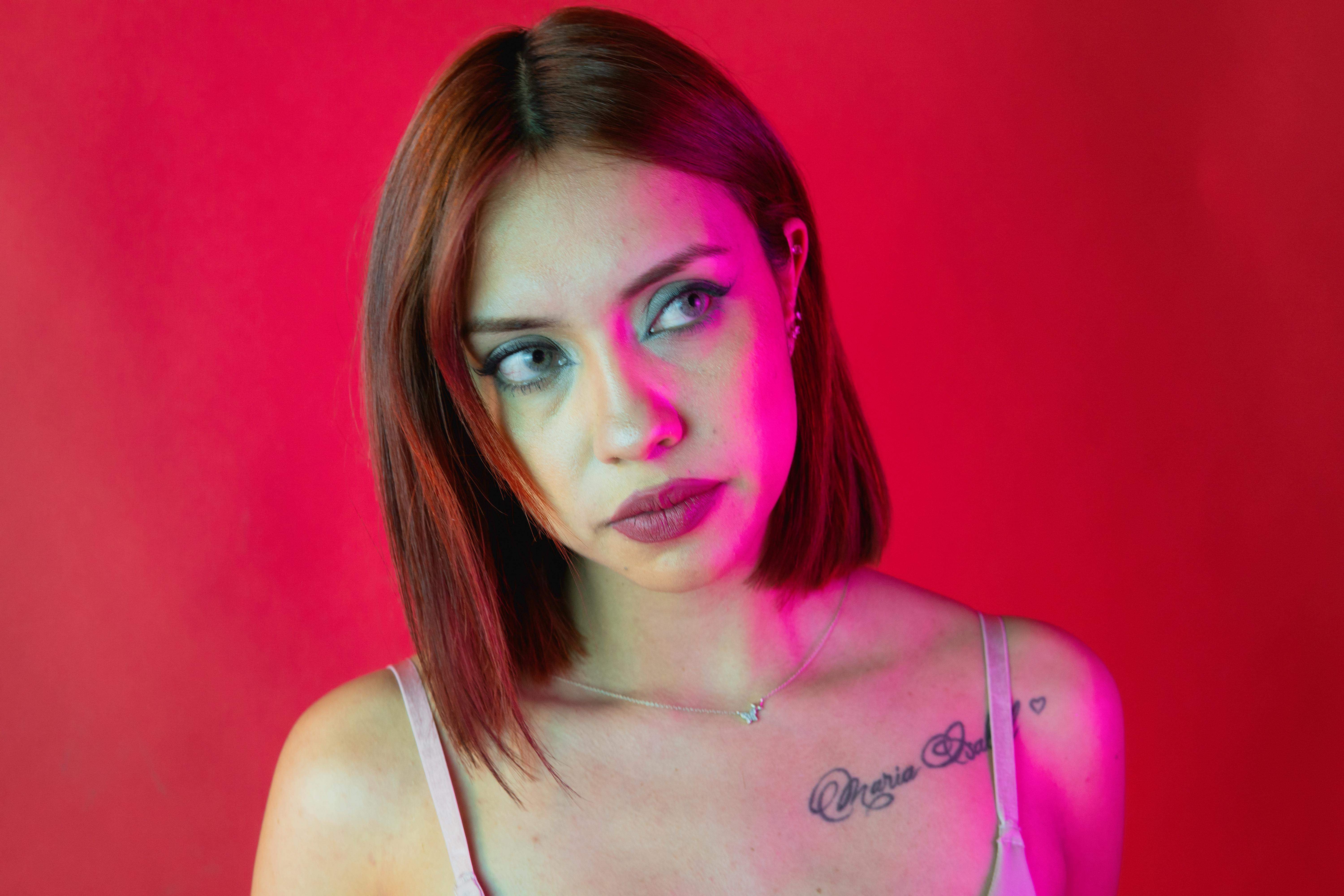 A striking portrait of a woman with red hair and a tattoo against a vibrant pink backdrop.
