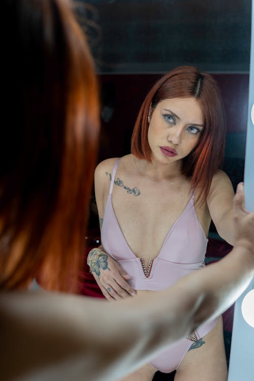 A woman with red hair in a pink bikini is looking in the mirror