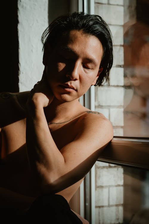 A shirtless man leaning against a window