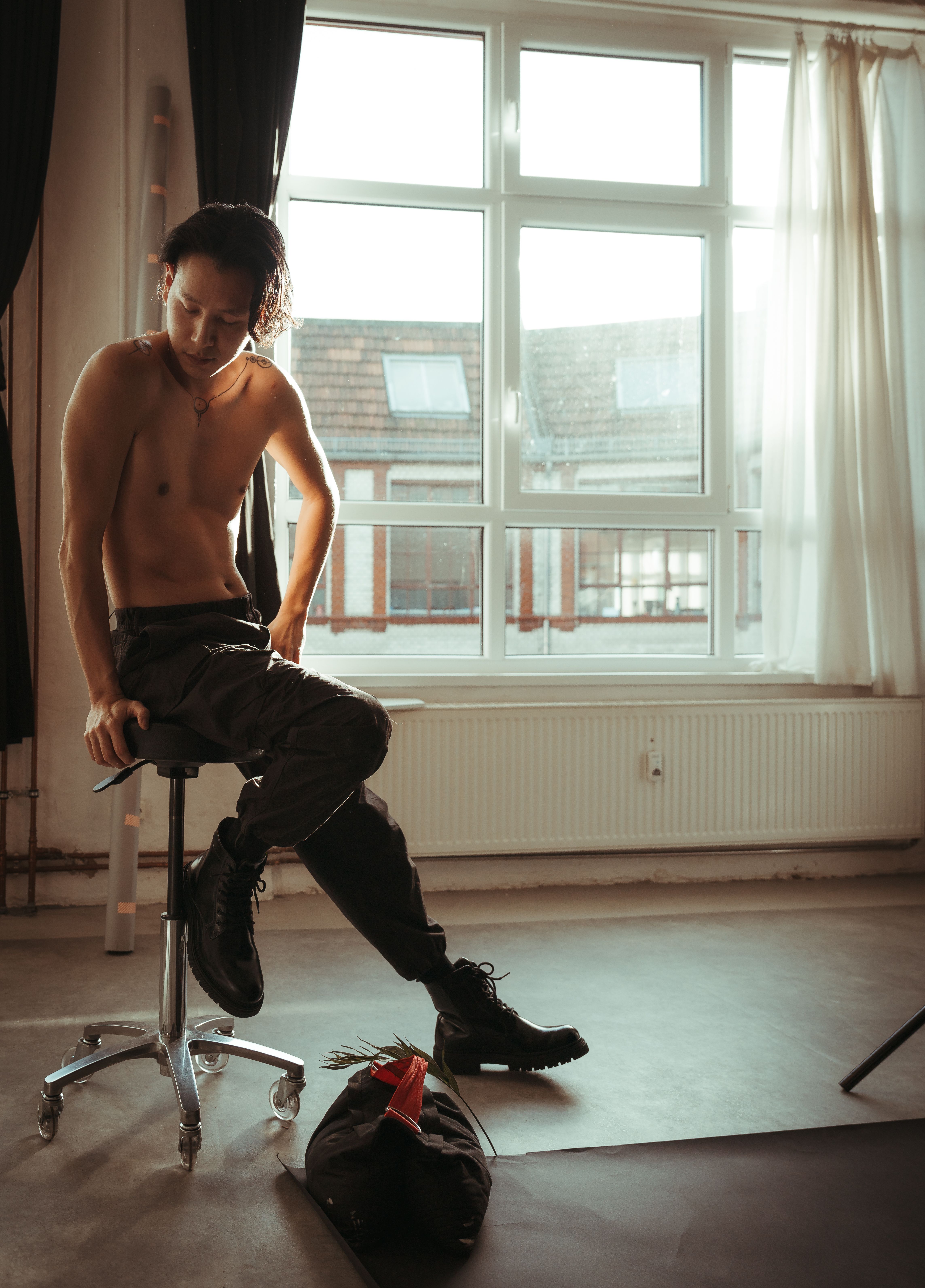 A shirtless man sitting on a chair by a large window, casting soft ambient light.