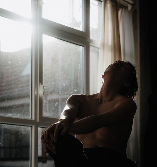 Free A shirtless man sits pensively by a sunlit window, capturing a serene indoor moment. Stock Photo