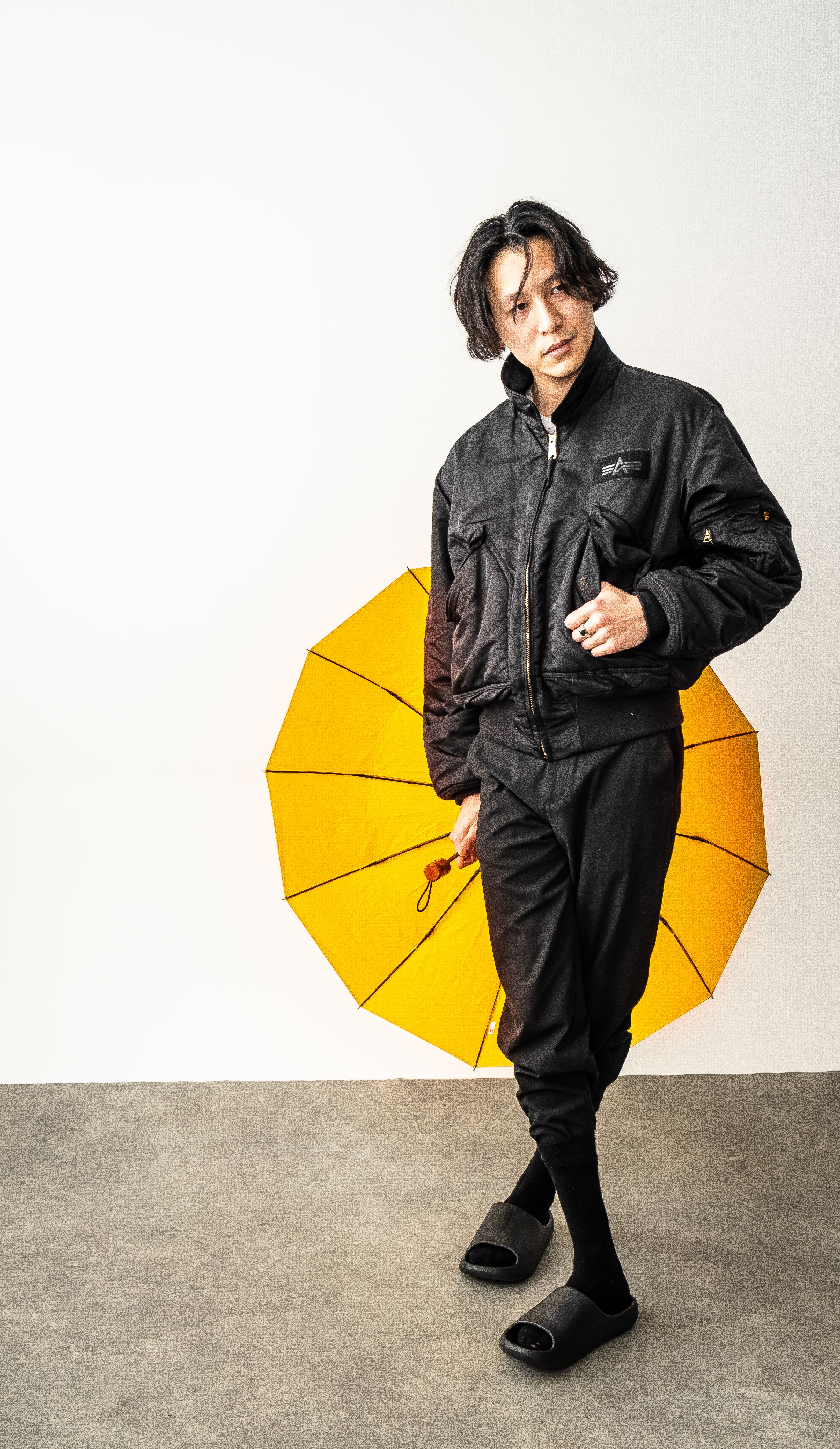 Asian man posing in black clothes with a vibrant yellow umbrella.