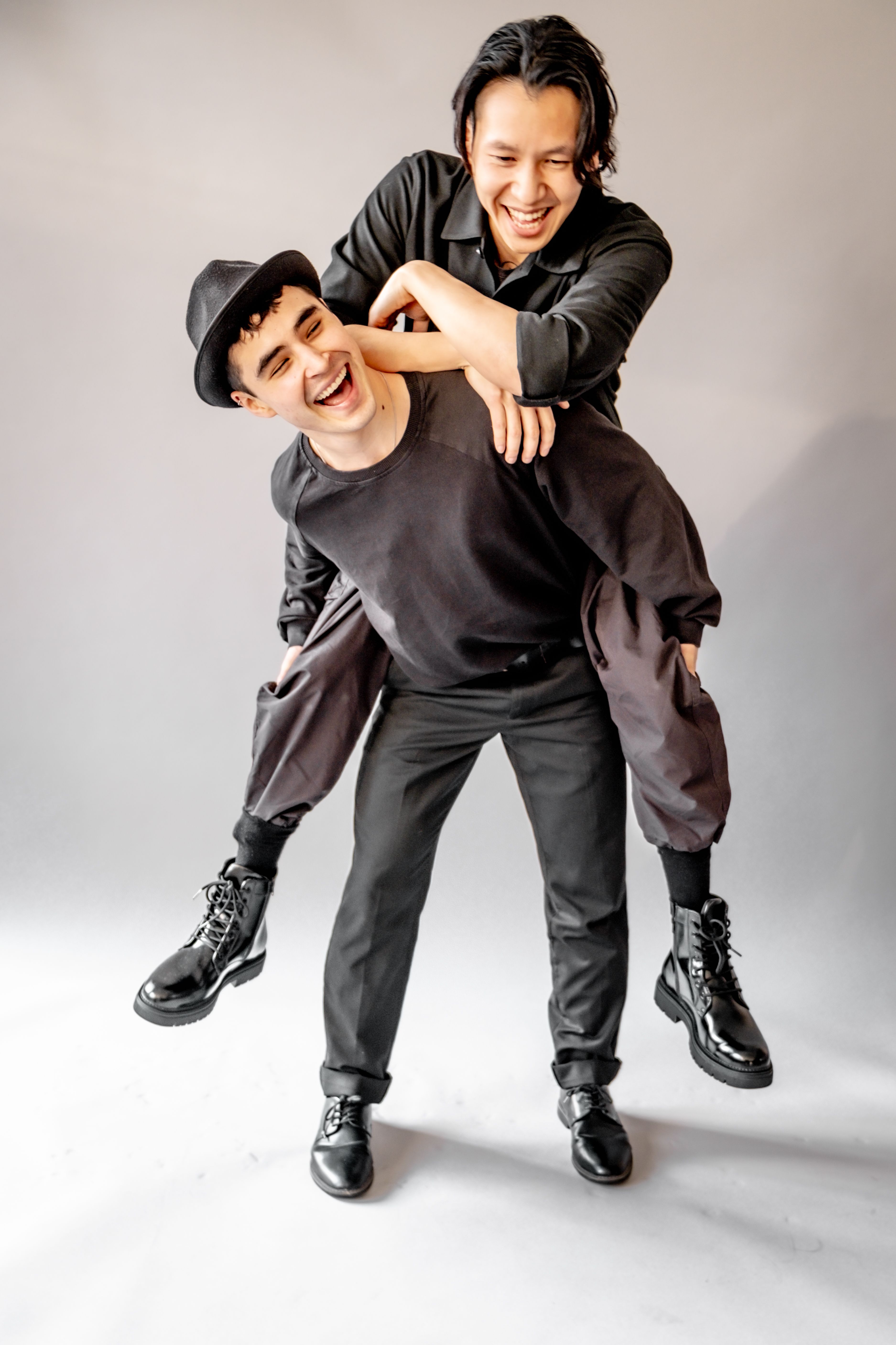 Two young men in black clothing playfully posing for a fashion portrait on a white background.