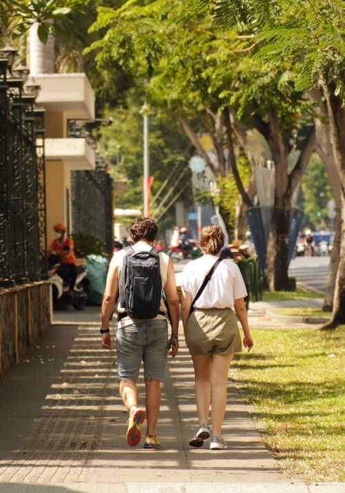 Free A couple holding hands strolling down a sunny city street surrounded by trees and buildings. Stock Photo