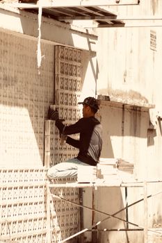 A construction worker lays tiles on a building facade under bright sunlight.