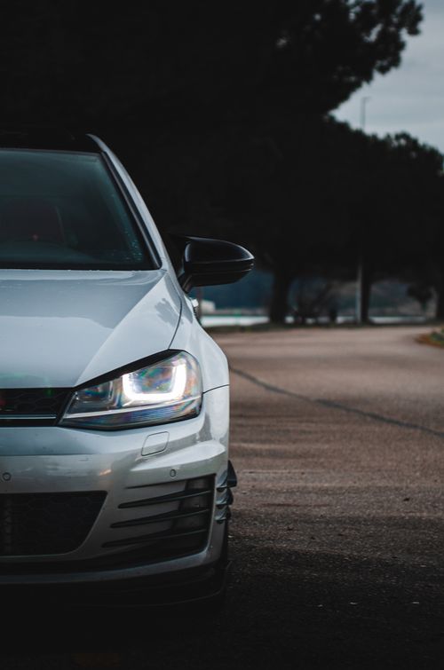 Free Dynamic half-body view of a white Volkswagen Golf GTI on a tree-lined outdoor road. Stock Photo