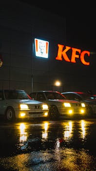 Rain-soaked street reflecting KFC neon sign with parked cars at night.
