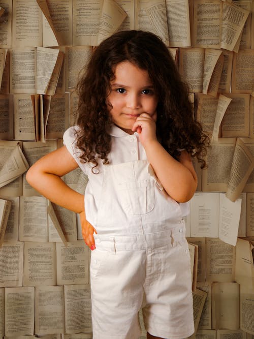 Free A little girl in white overalls standing in front of a wall of books Stock Photo
