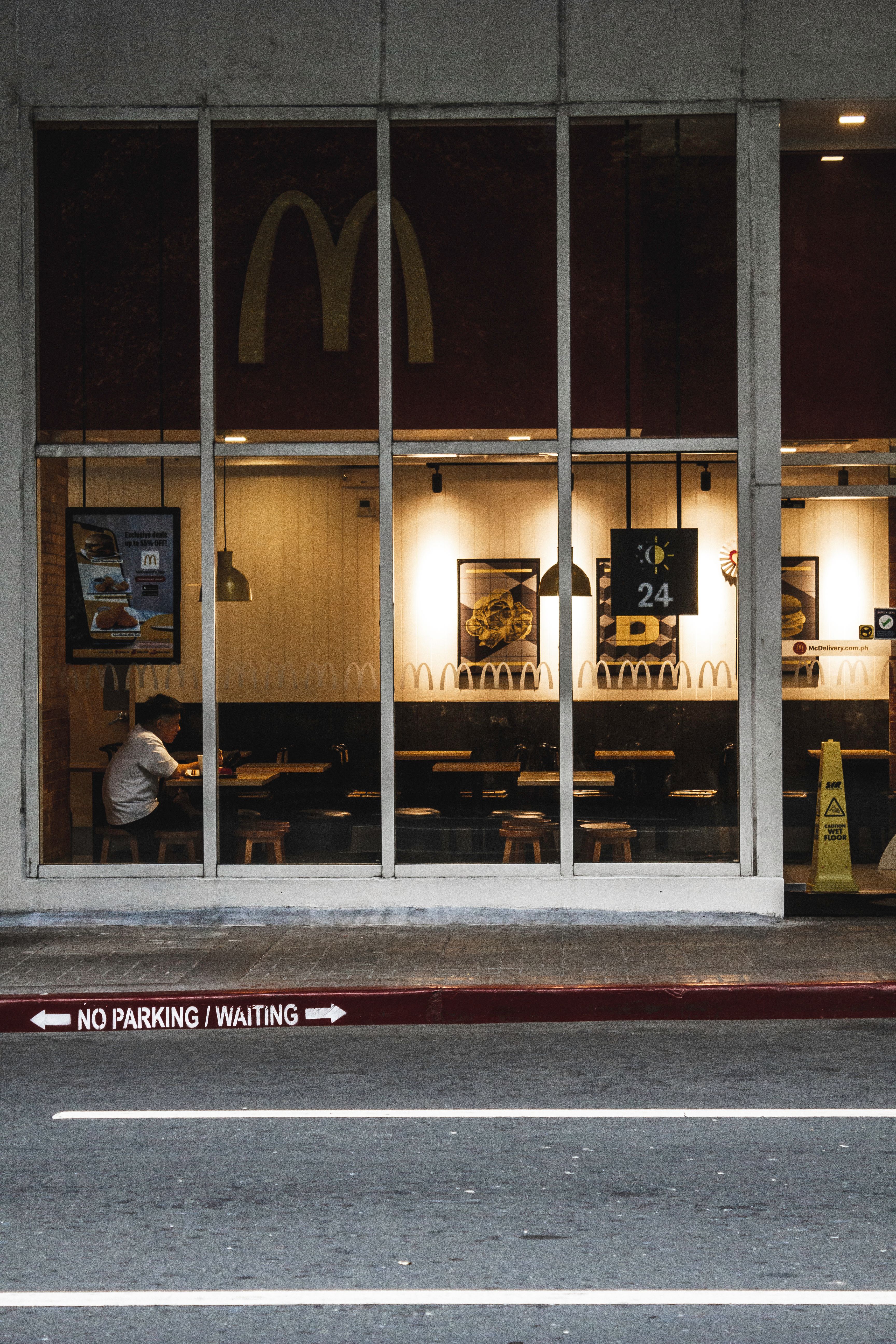 A solitary customer inside a well-lit fast food restaurant seen through large windows at night.