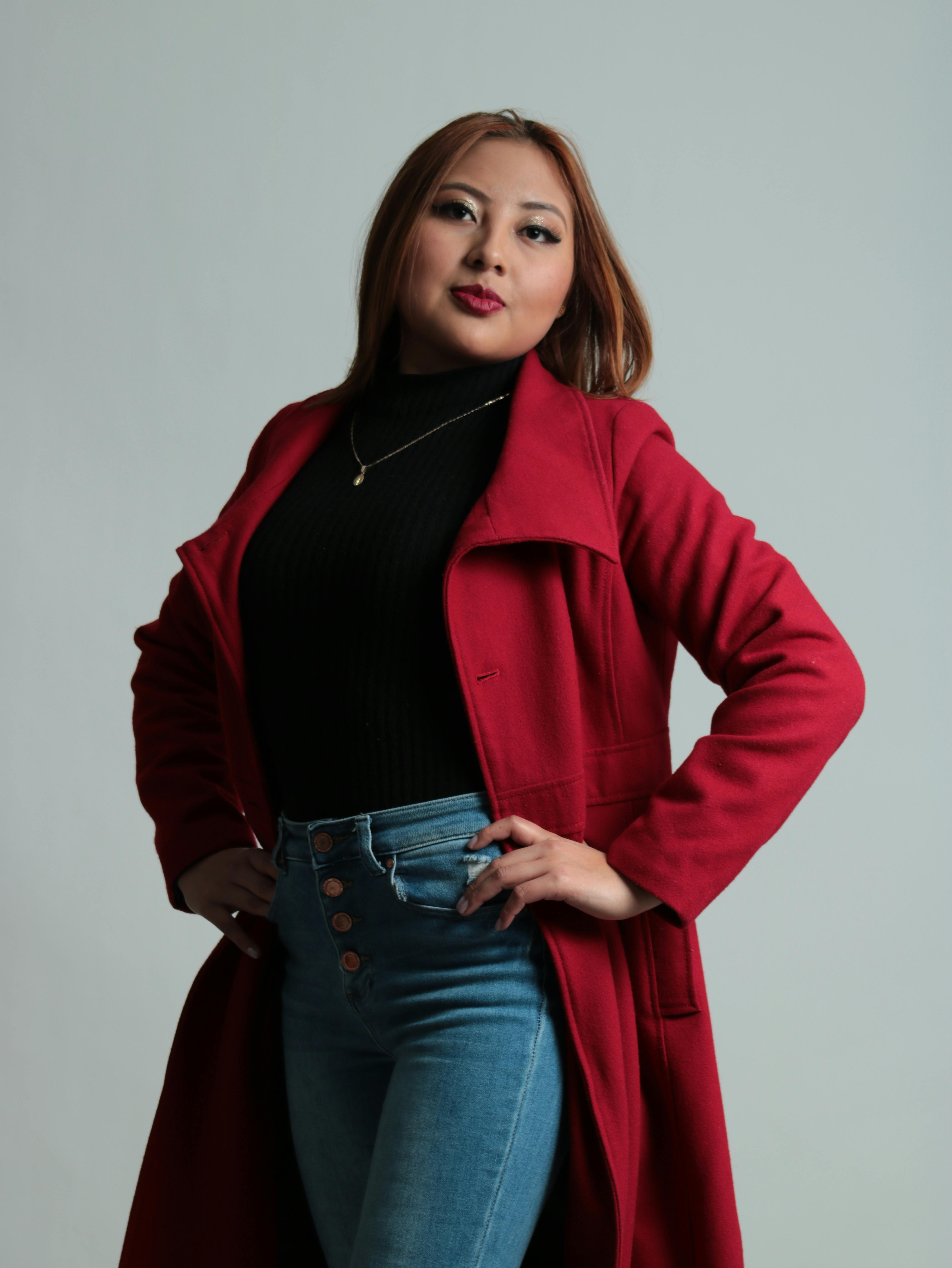 Confident woman posing in a red coat and jeans against a white background.
