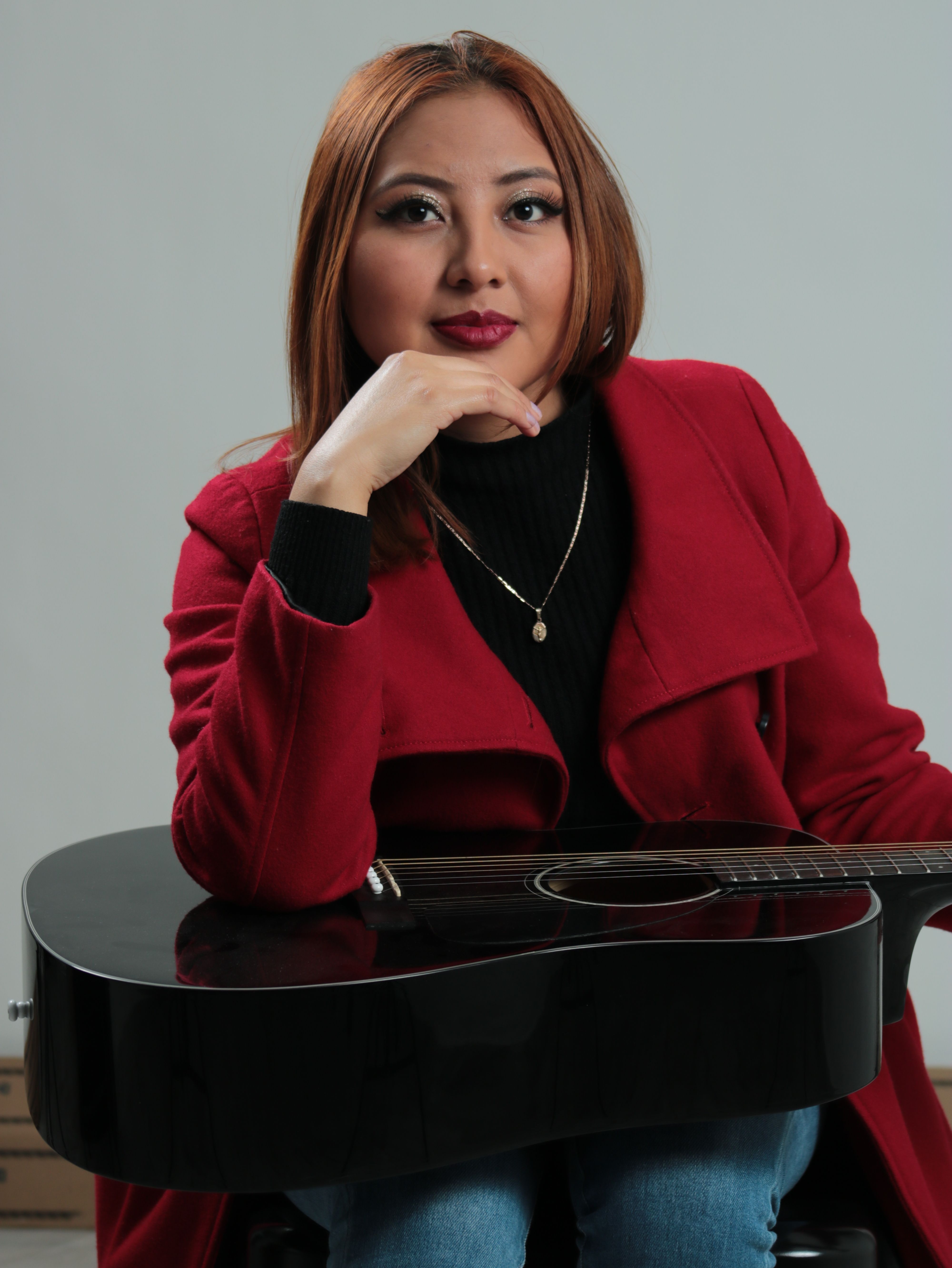 A stylish woman in a red coat poses with a guitar in a studio setting.