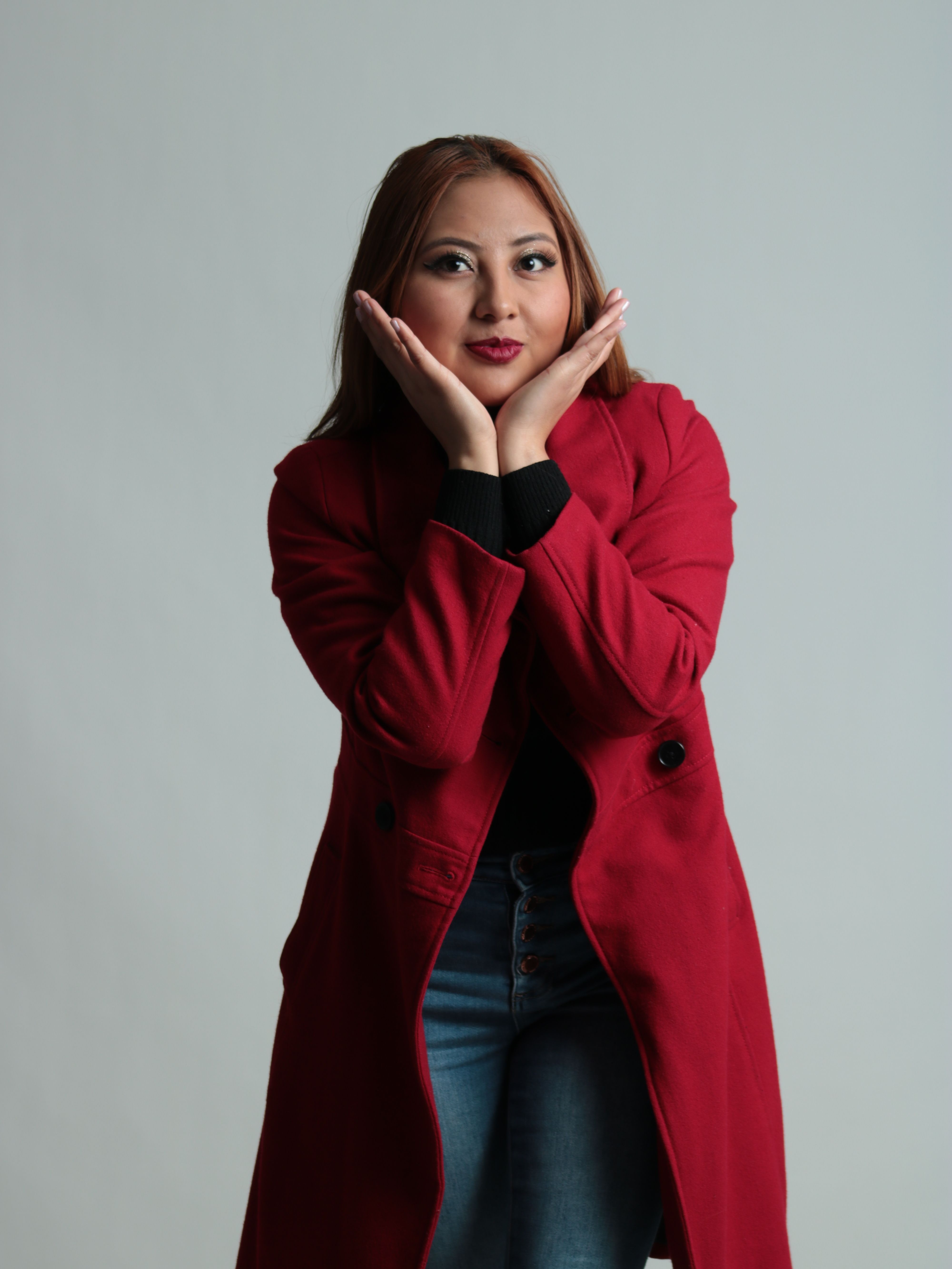 Studio portrait of a woman posing in a red coat with hands on cheeks against a gray background.