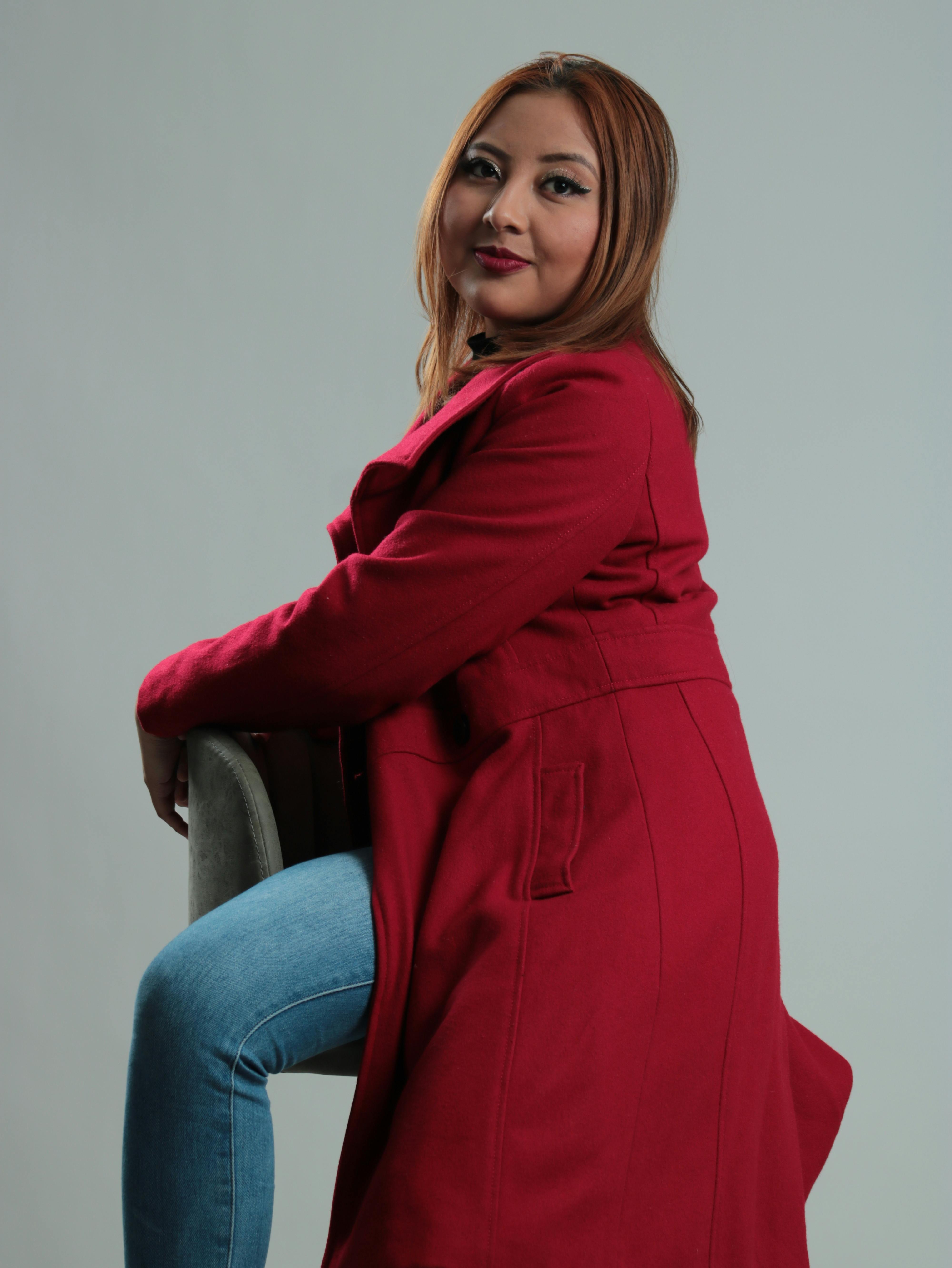 A stylish woman wearing a red coat and jeans sits gracefully in a studio setting. Modern fashion photography.