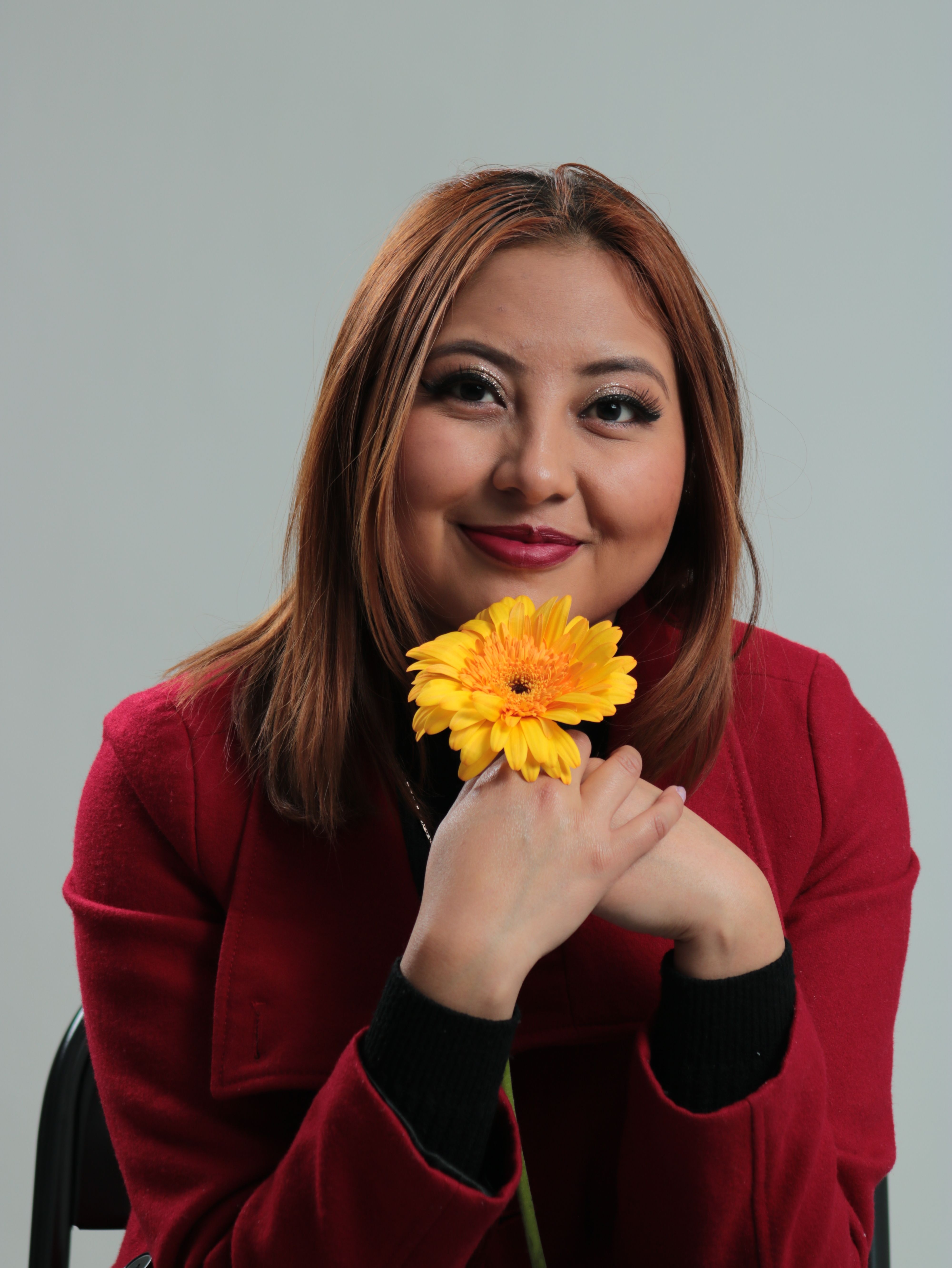 Smiling woman in a red jacket holding a yellow flower in a studio setting.