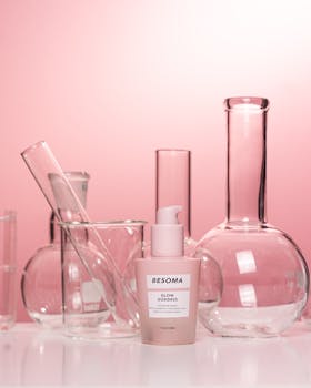 Cosmetic bottle surrounded by scientific glassware against a pink background, studio setting.