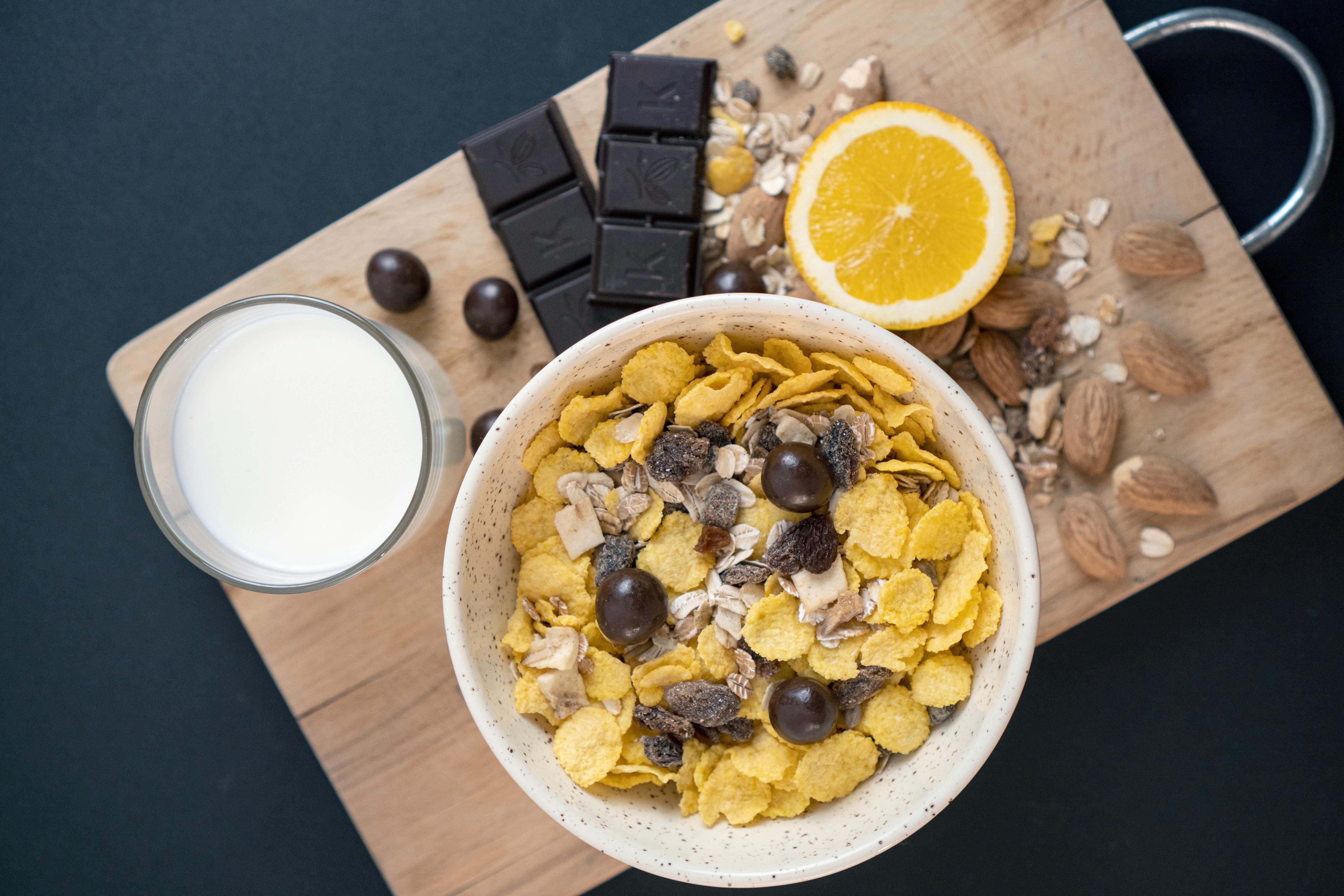 A close-up of a nutritious breakfast featuring cereal, chocolate, orange, and milk on a wooden board.