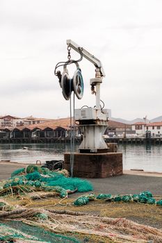 An industrial crane on a harbor with fishing nets and distant town buildings under a cloudy sky.