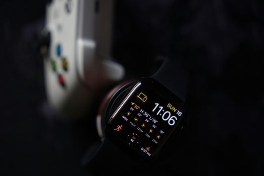 Close-up view of a smartwatch displaying time and weather next to a game controller on a dark background.