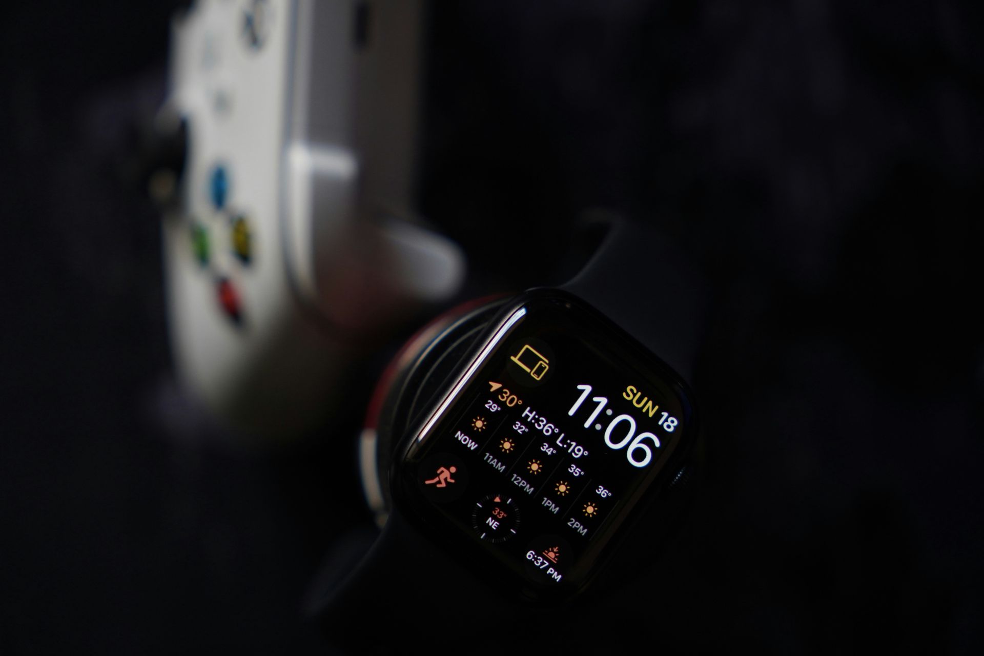 Close-up view of a smartwatch displaying time and weather next to a game controller on a dark background.