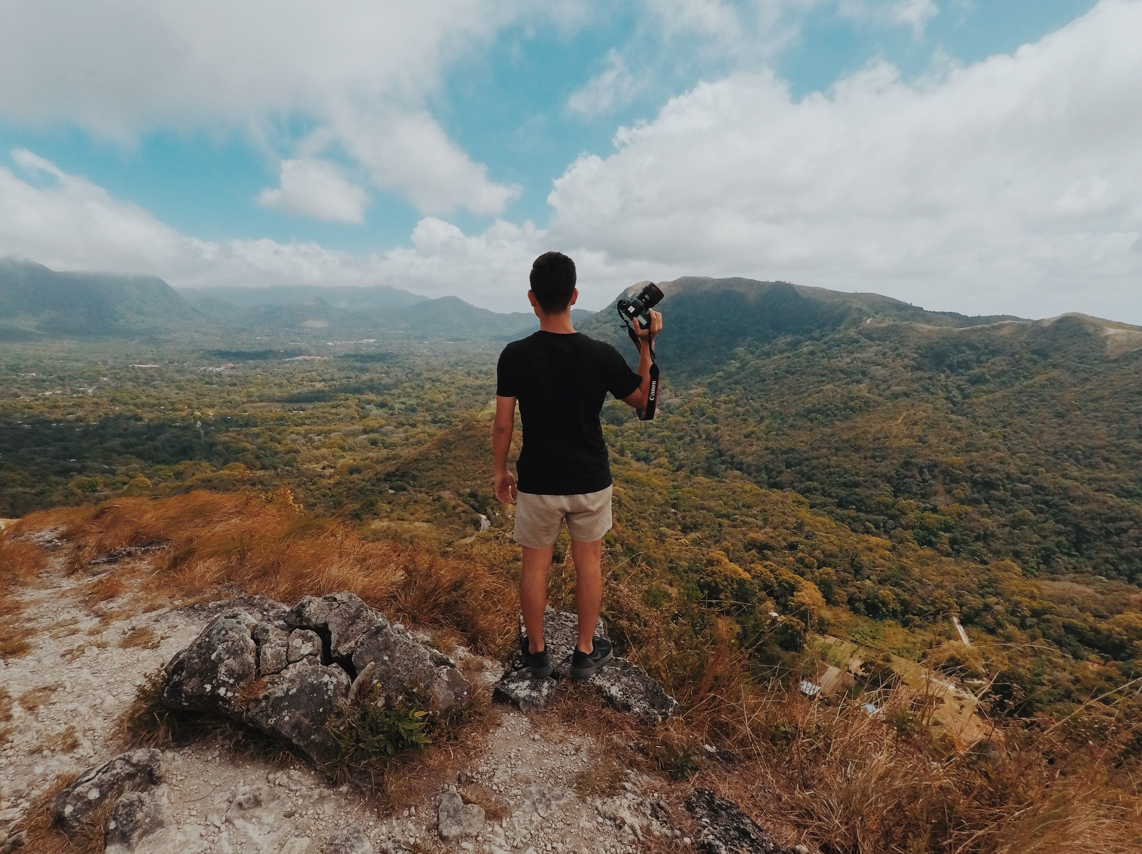 A man holding a camera stands on a rocky hill, gazing at a vast, scenic mountain landscape under a cloudy sky.