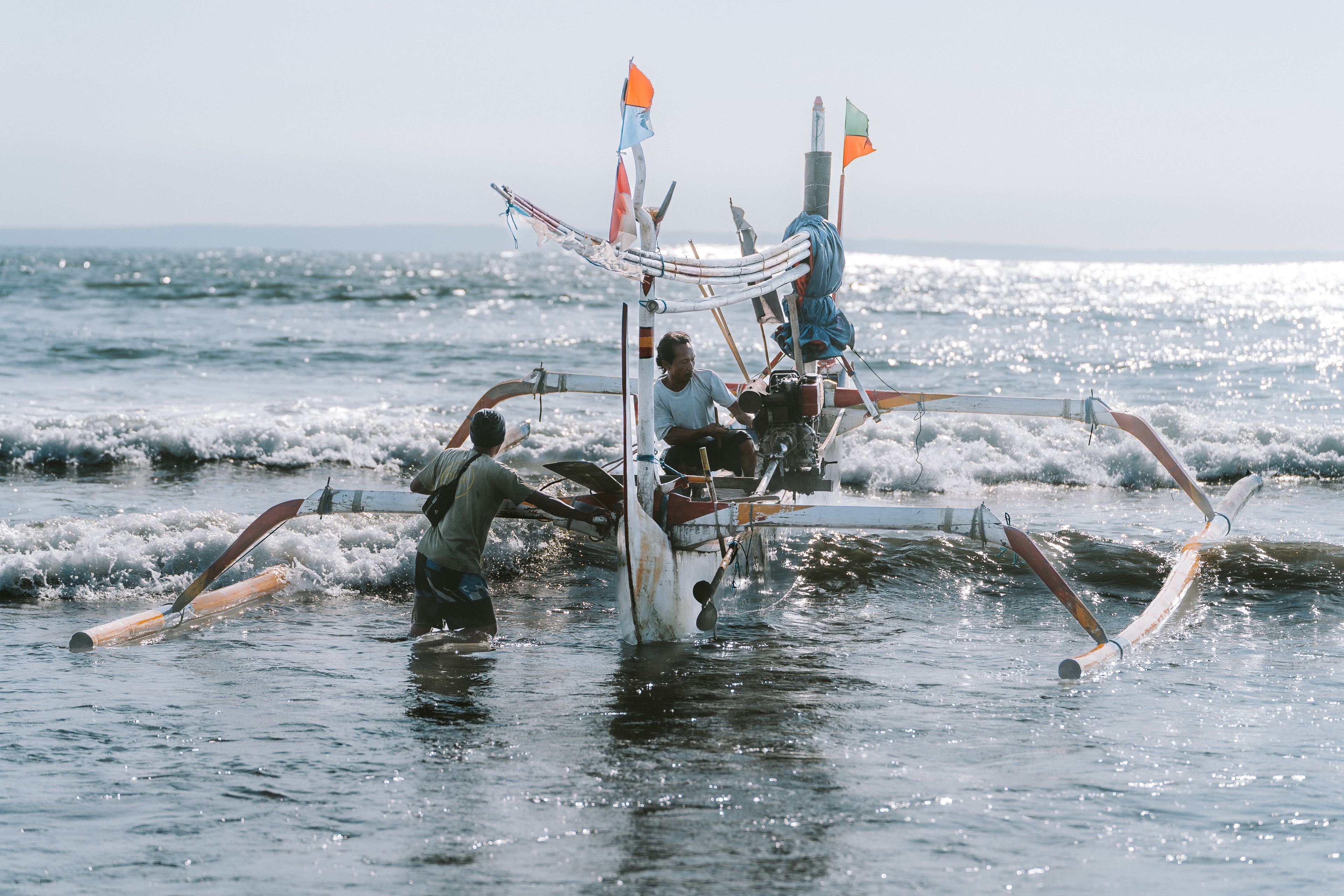 Fishermen launching a traditional boat in Pulukan, Bali, with scenic ocean waves in the background.