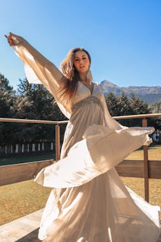 Woman in flowing beige dress poses gracefully under summer sunlight on a scenic balcony.