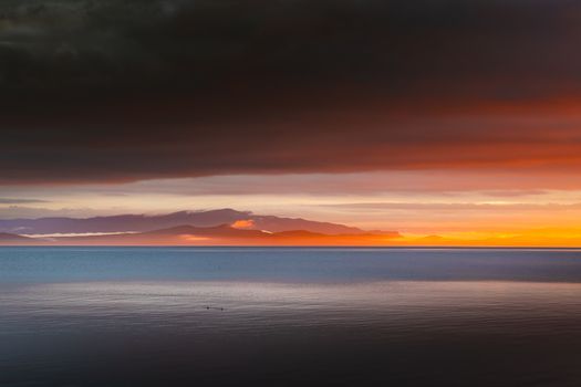 A stunning seascape capturing a dramatic sunset over the ocean with illuminated clouds and a distant mountain silhouette.