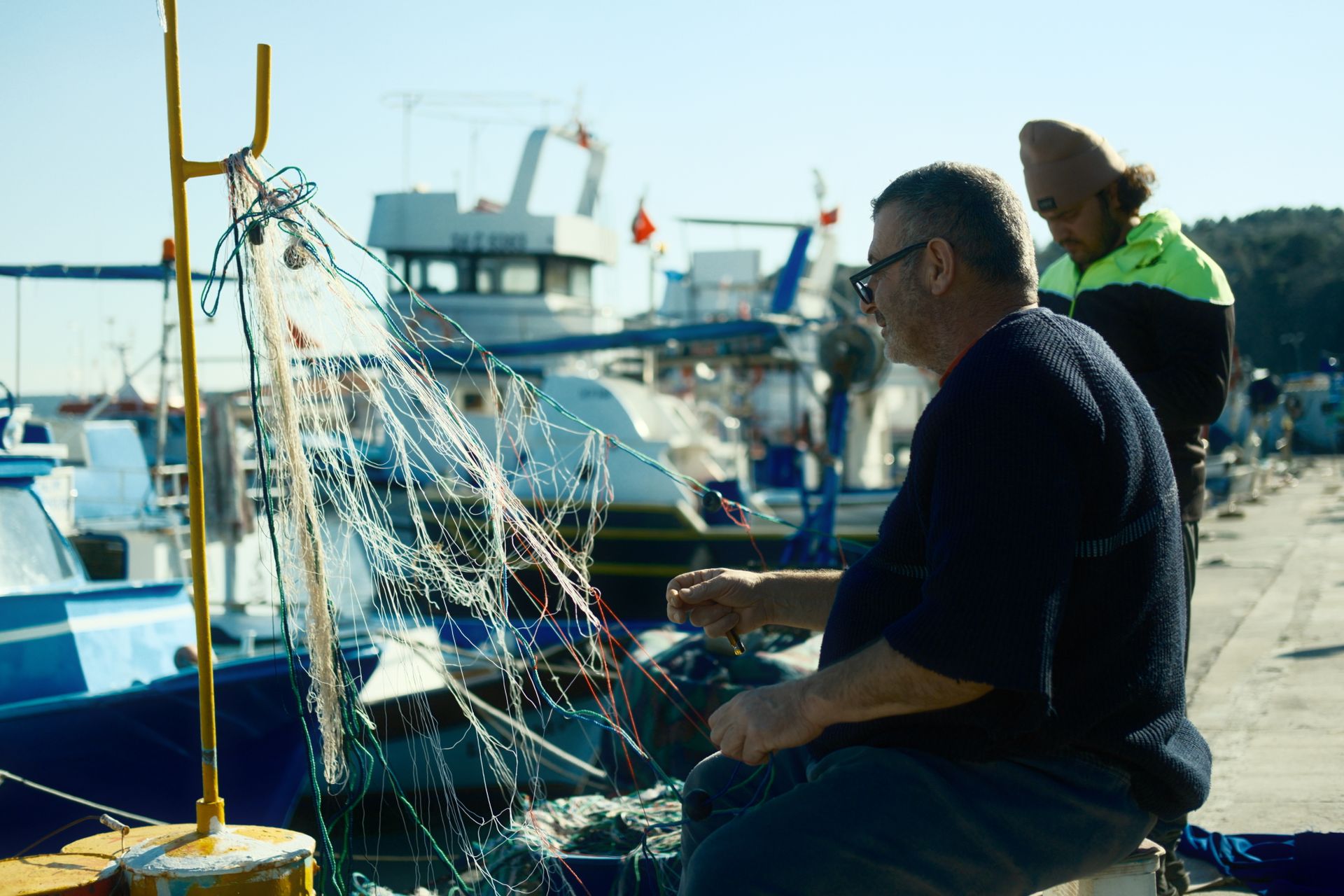 Two fishermen mending nets by the harbor with boats moored nearby, showcasing daily maritime life.