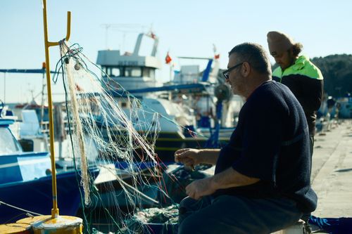 Free Two fishermen mending nets by the harbor with boats moored nearby, showcasing daily maritime life. Stock Photo