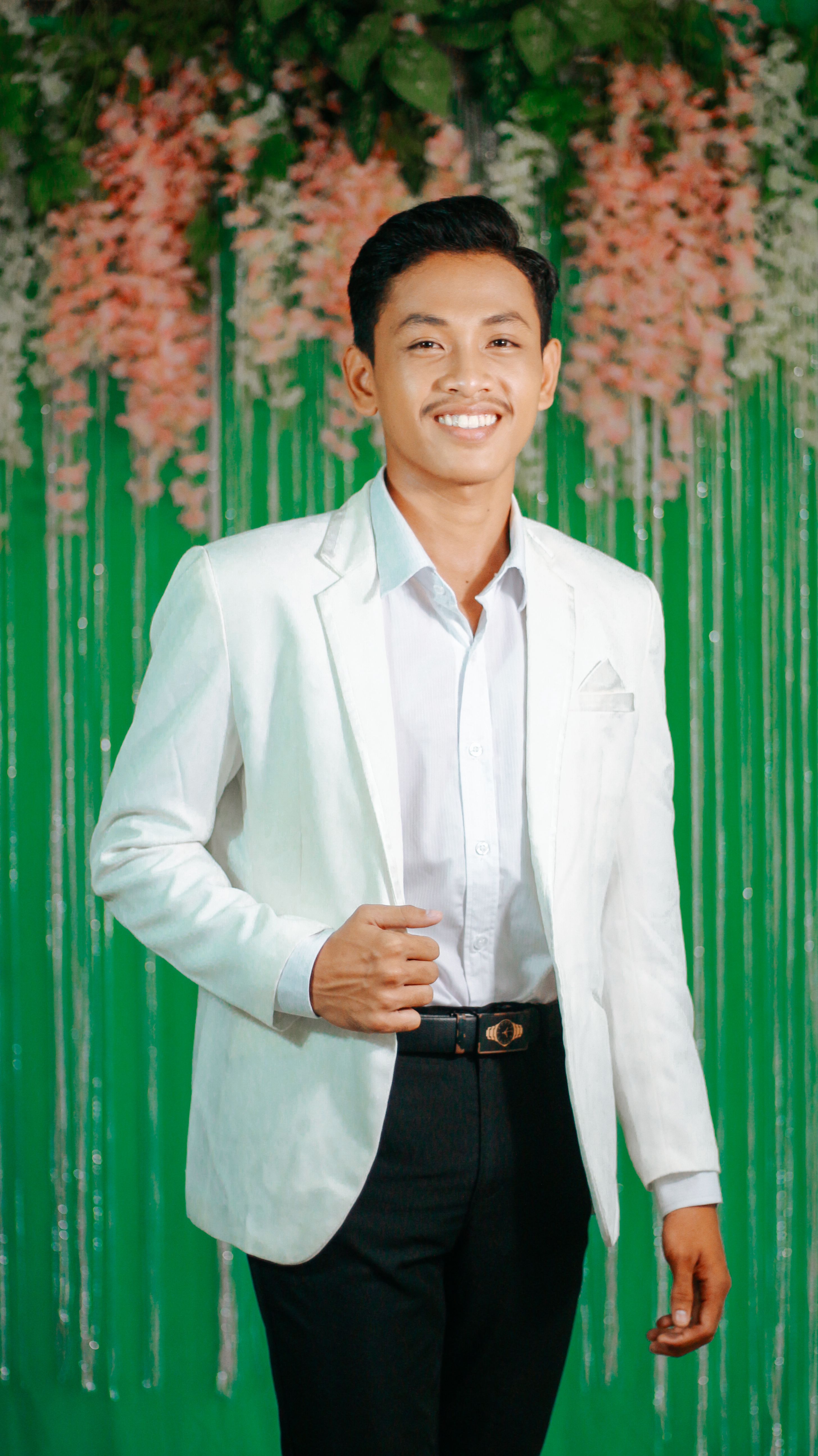 Confident young man in a white suit smiling against a floral backdrop.