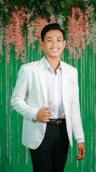 Confident young man in a white suit smiling against a floral backdrop.