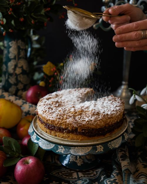 Free Hands sprinkling powdered sugar on a homemade cake surrounded by fruit and floral decor. Stock Photo