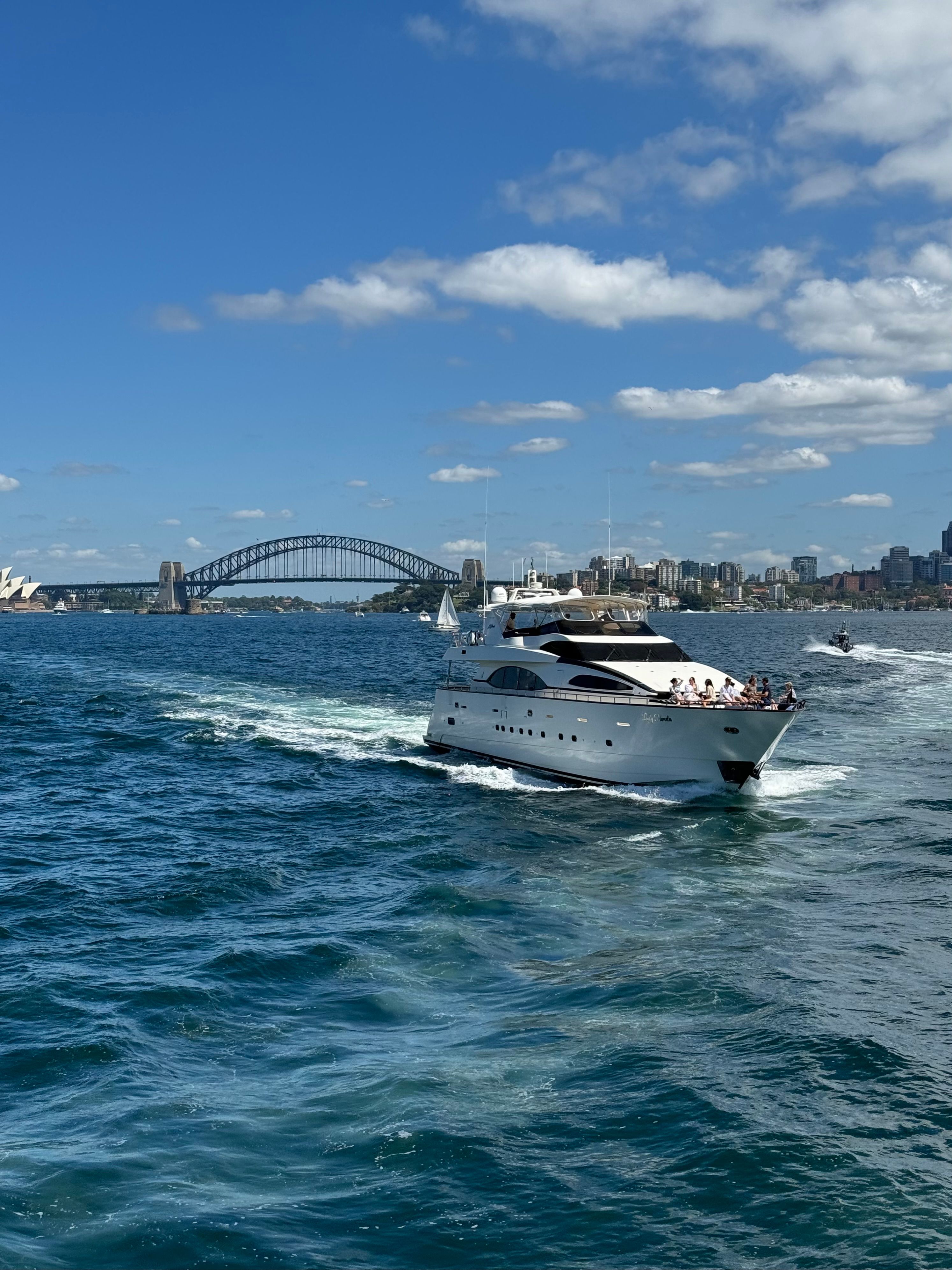 A luxury yacht sails in Sydney Harbor, with the iconic Sydney Harbour Bridge in the background on a sunny day.