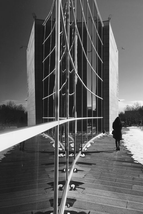 Free Monochromatic photo of a person walking by a modern glass building reflecting symmetry. Stock Photo