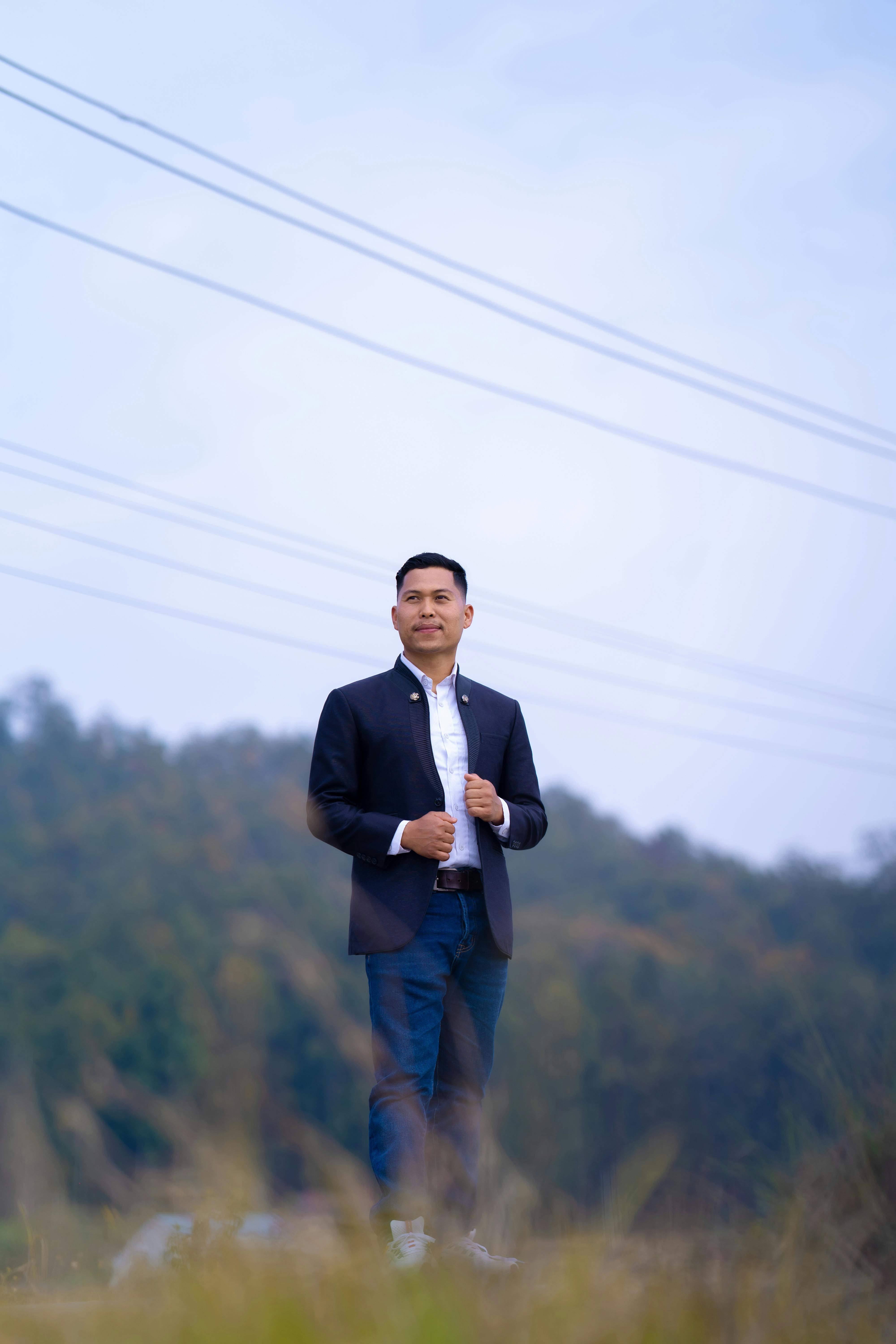 Asian man wearing a dinner jacket and jeans, standing in an open field, showcasing fashion with confidence.