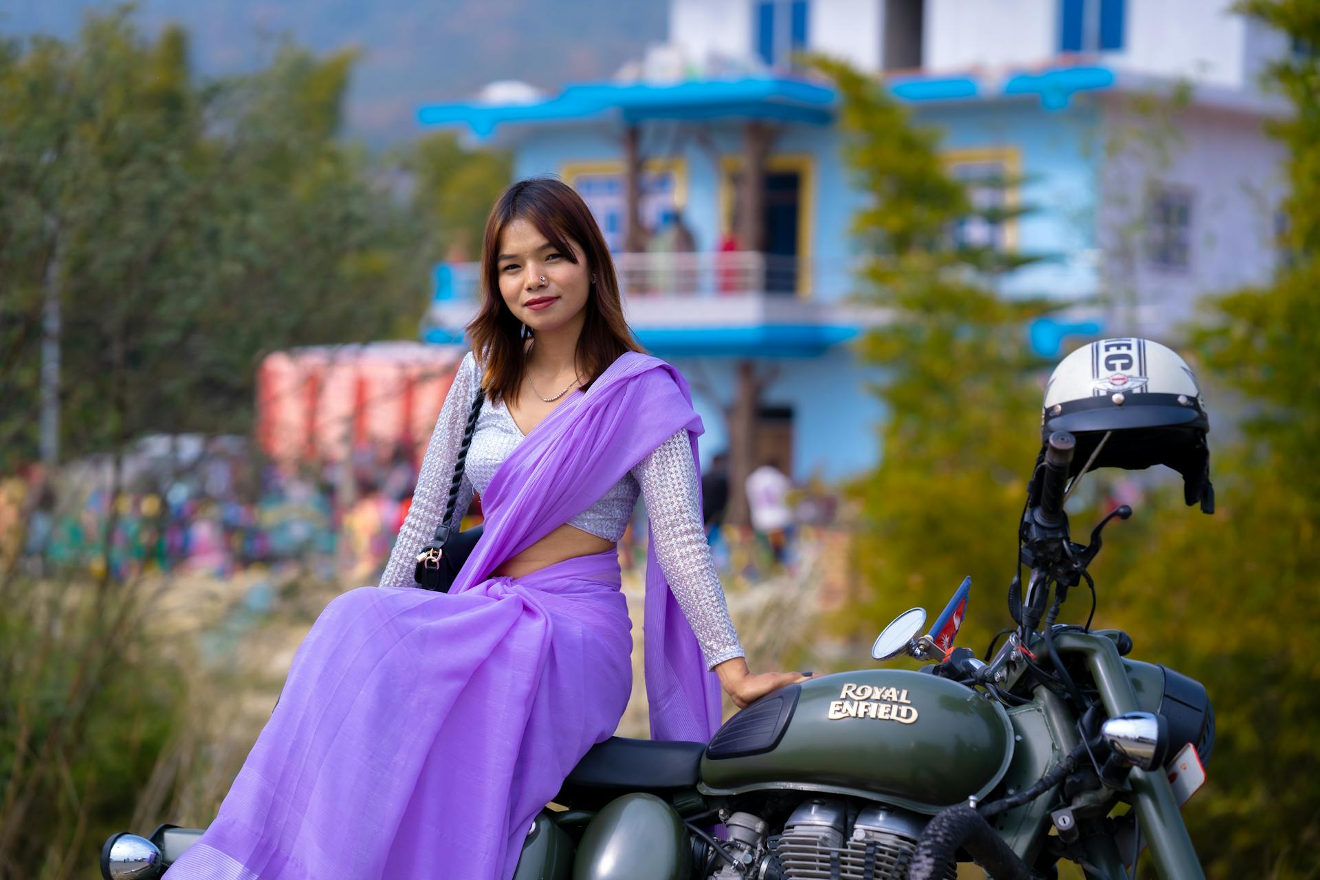 A fashionable woman in a purple sari posing confidently on a Royal Enfield motorcycle outdoors.