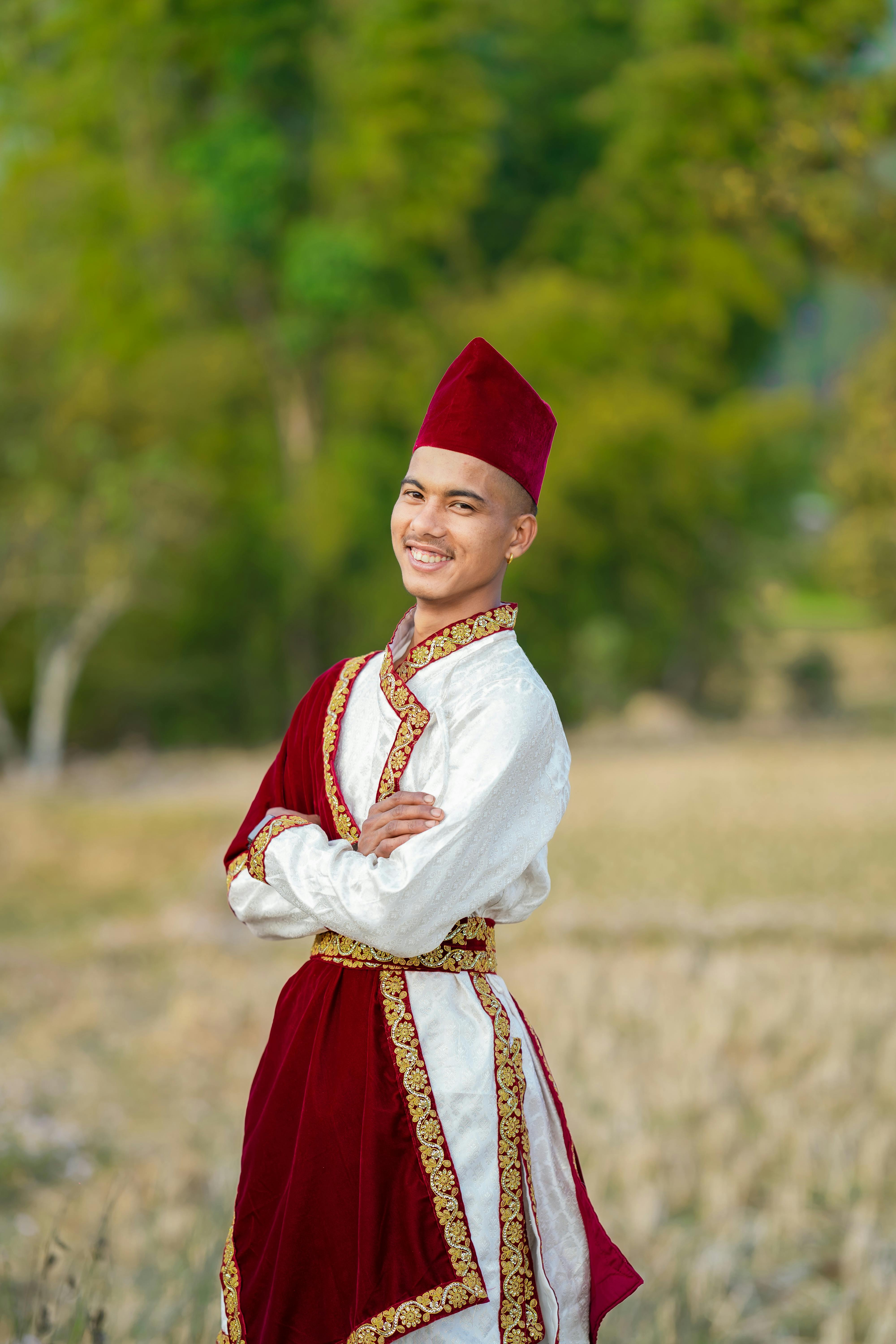 Smiling man in traditional red and white attire standing in a vibrant green outdoor scene.