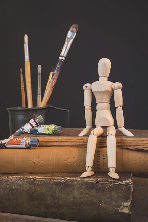 A wooden doll sits on top of books and paint brushes
