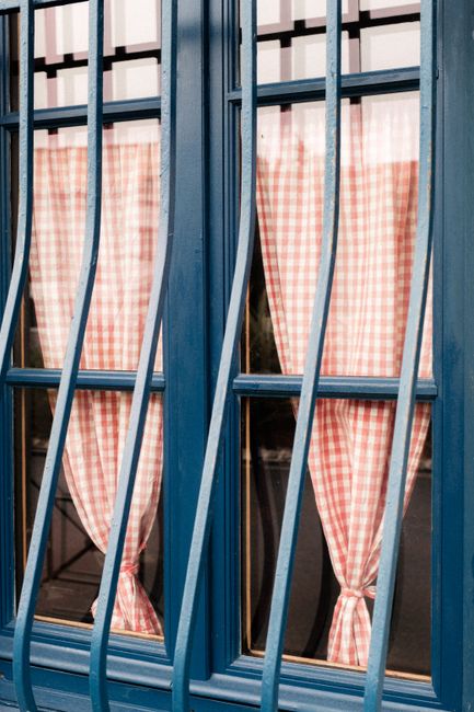 Blue window with pink gingham curtains in Biarritz, France, exudes rustic charm.