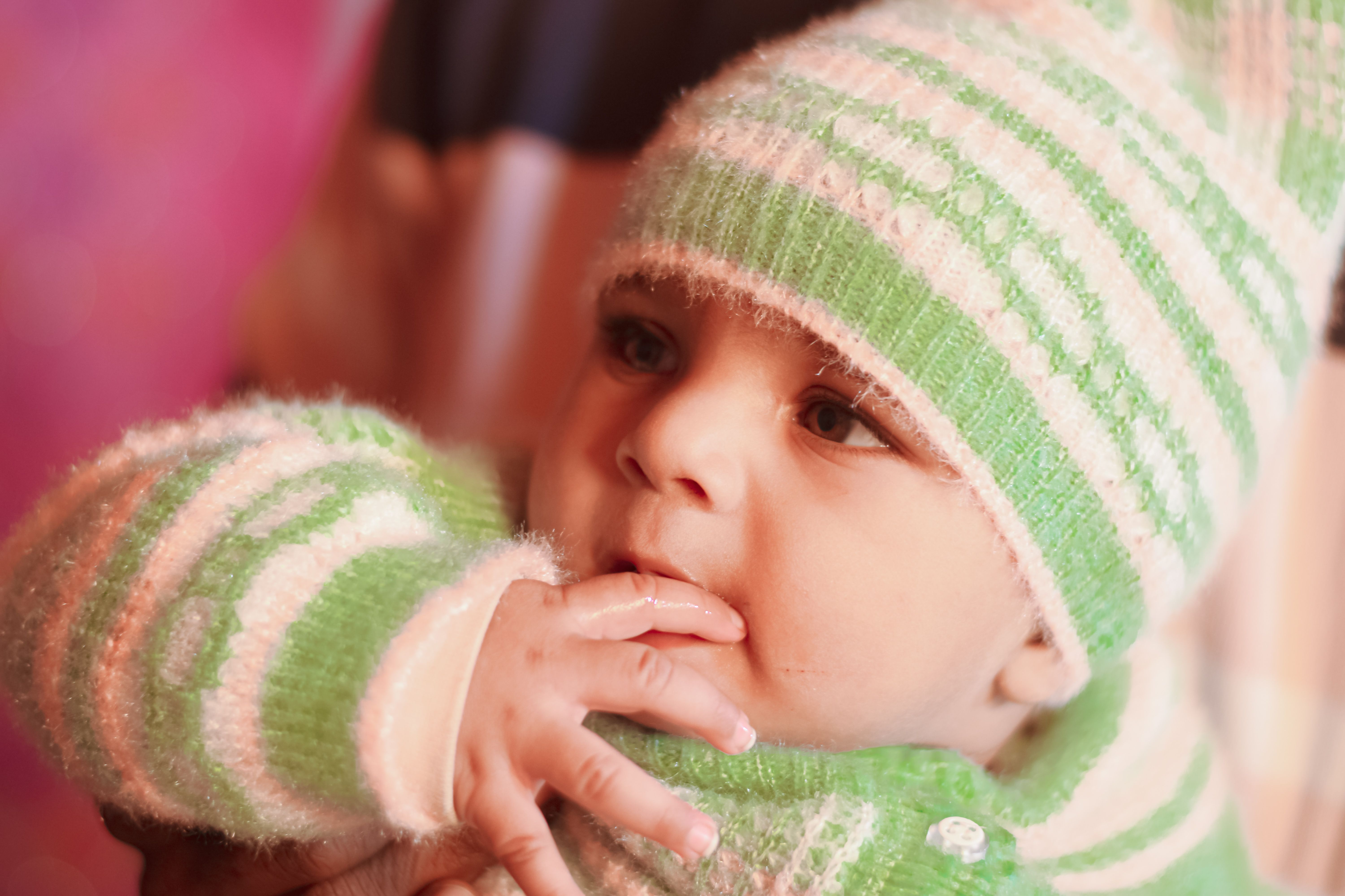 A close-up portrait of a baby in a green woolen hat and sweater, with fingers in mouth.