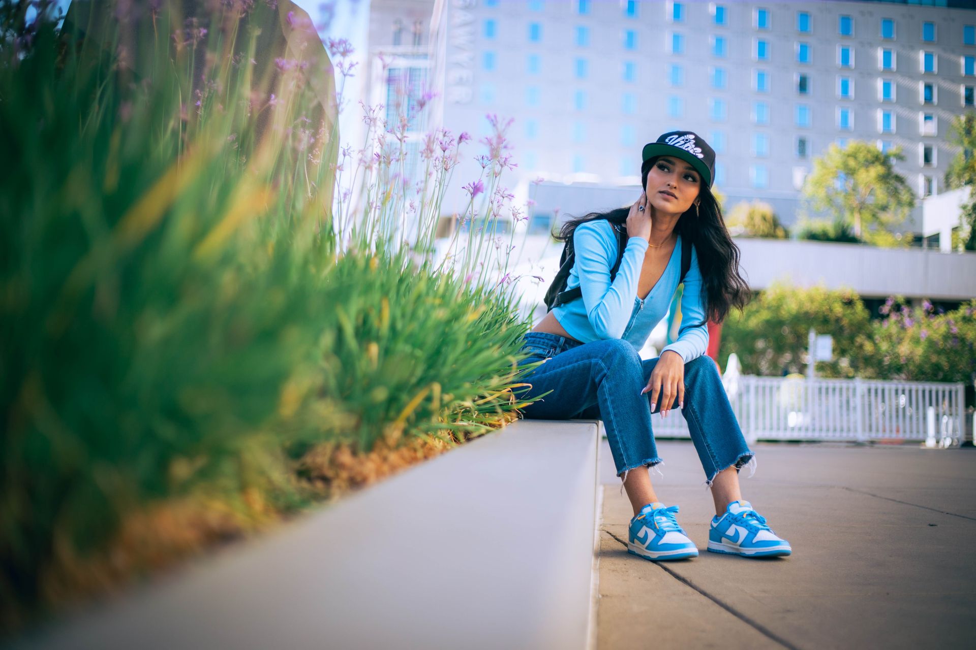Woman in urban setting wearing blue jeans and top with black cap, seated amid flowers.